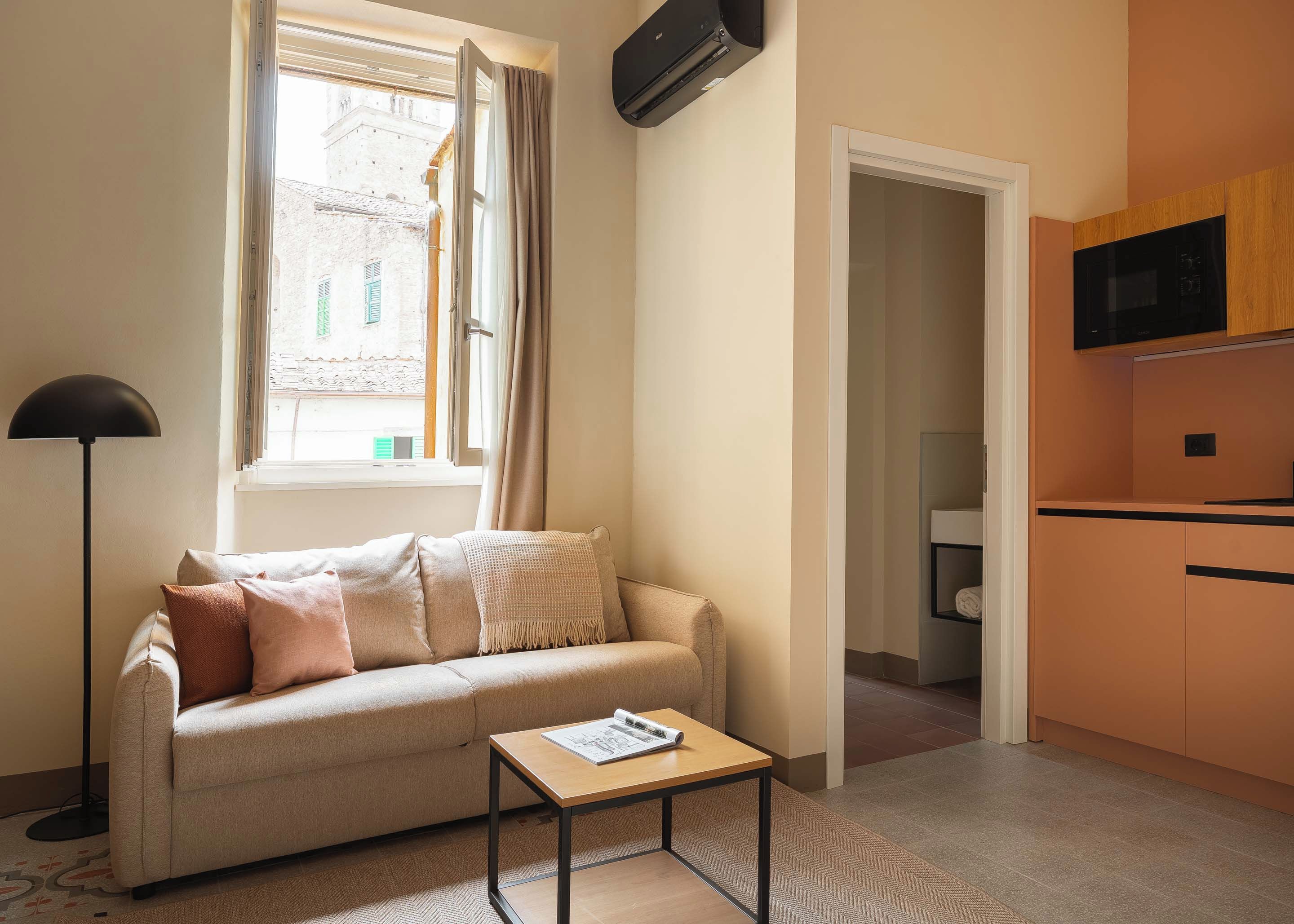 A cozy living space with a sofa, a small table, a window with curtains, and a kitchenette area.