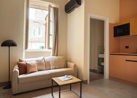 Un espacio de sala cómodo con un sofá, una mesita, una ventana con cortinas y una zona de kitchenette.