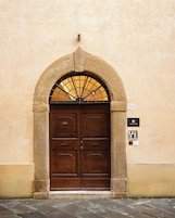 Una puerta doble de madera con arco superior y una celosía metálica, ubicada en una pared color beige con una placa de número arriba.