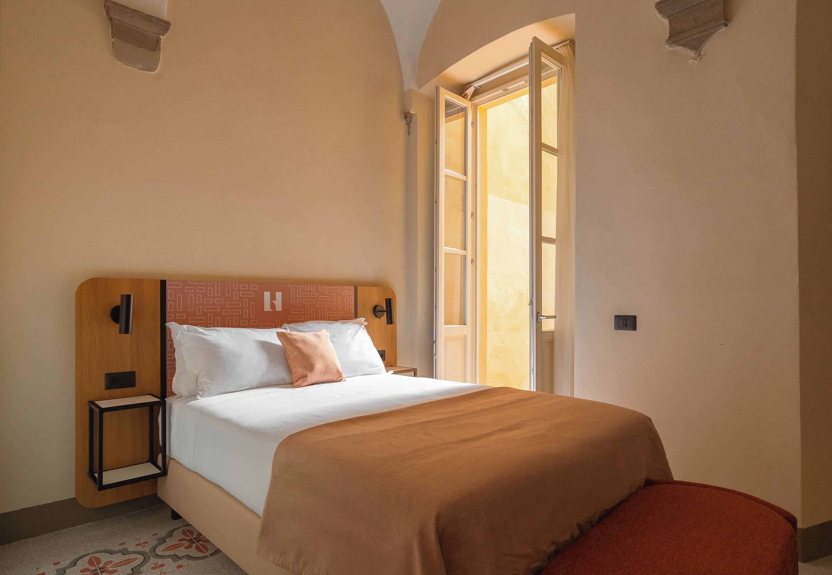 Una habitación de hotel con una cama, mesita de noche y ventana con cortinas.