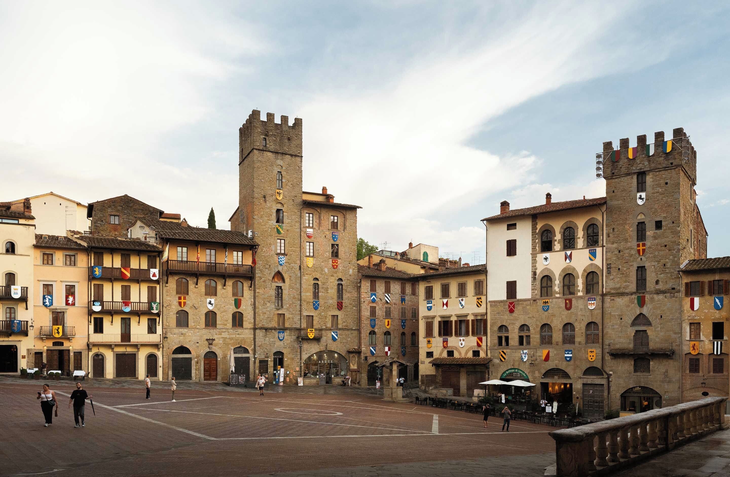 Una piazza storica con una torre imponente e numerosi edifici decorati con diversi stemmi araldici.