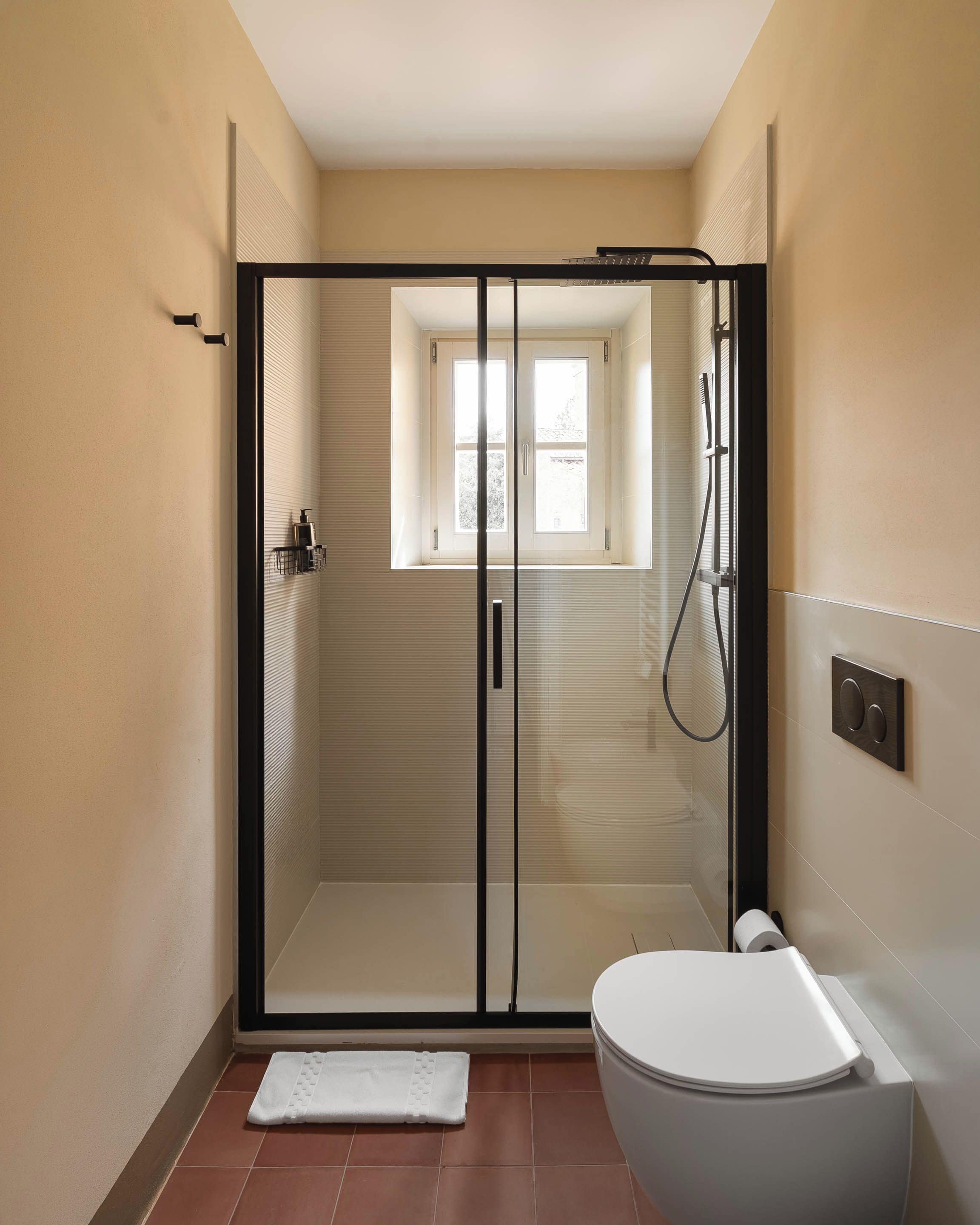 A modern bathroom with a glass-enclosed shower, a toilet, and a small window.