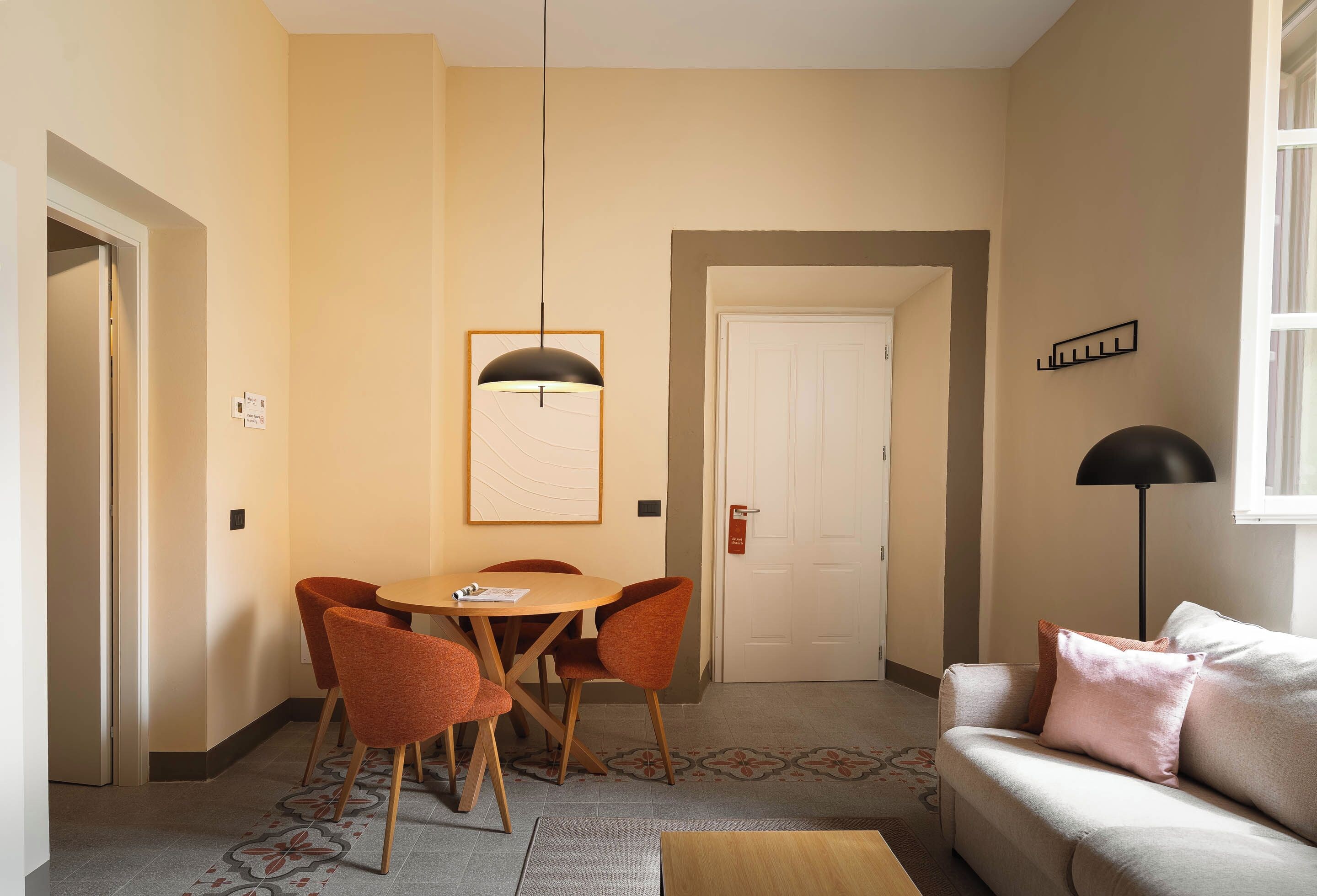 A hotel room with a dining area featuring a round table and four chairs, a sofa with cushions, a standing lamp, and a door with a sign.
