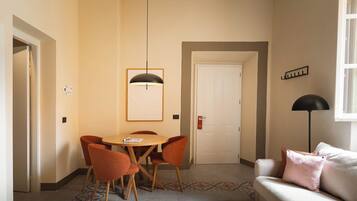 A hotel room with a dining area featuring a round table and four chairs, a sofa with cushions, a standing lamp, and a door with a sign.