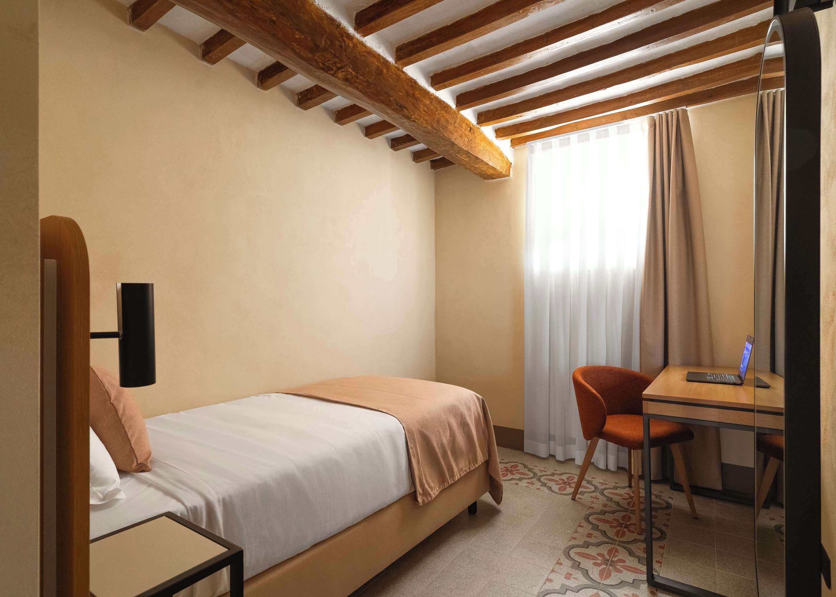 Een hotelkamer met een bed, een bureau met een laptop, een stoel en een raam met gordijnen.