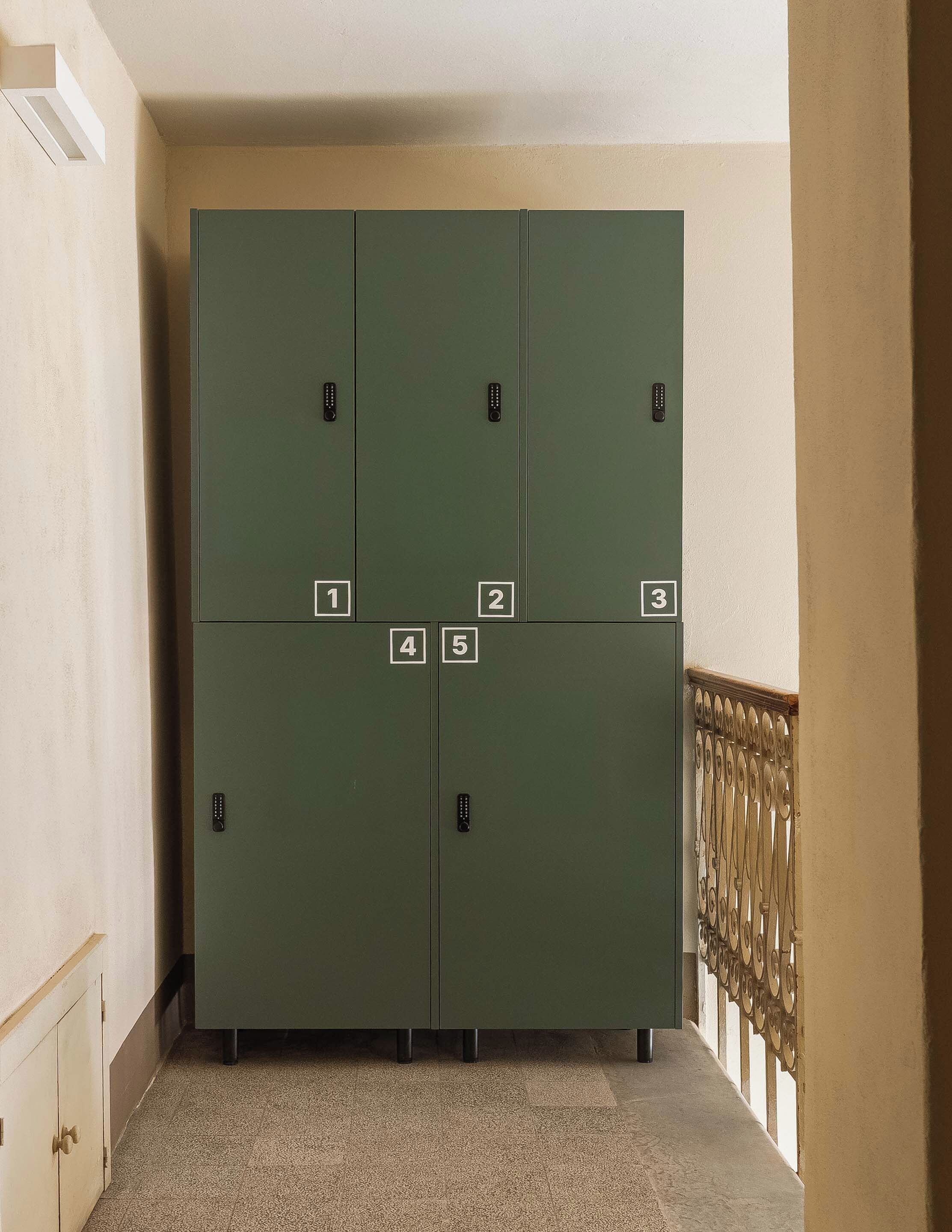 Eine Reihe von grünen, nummerierten Schließfächern von 1 bis 5, mit einem Metallgeländer und einer beigen Wand im Hintergrund.