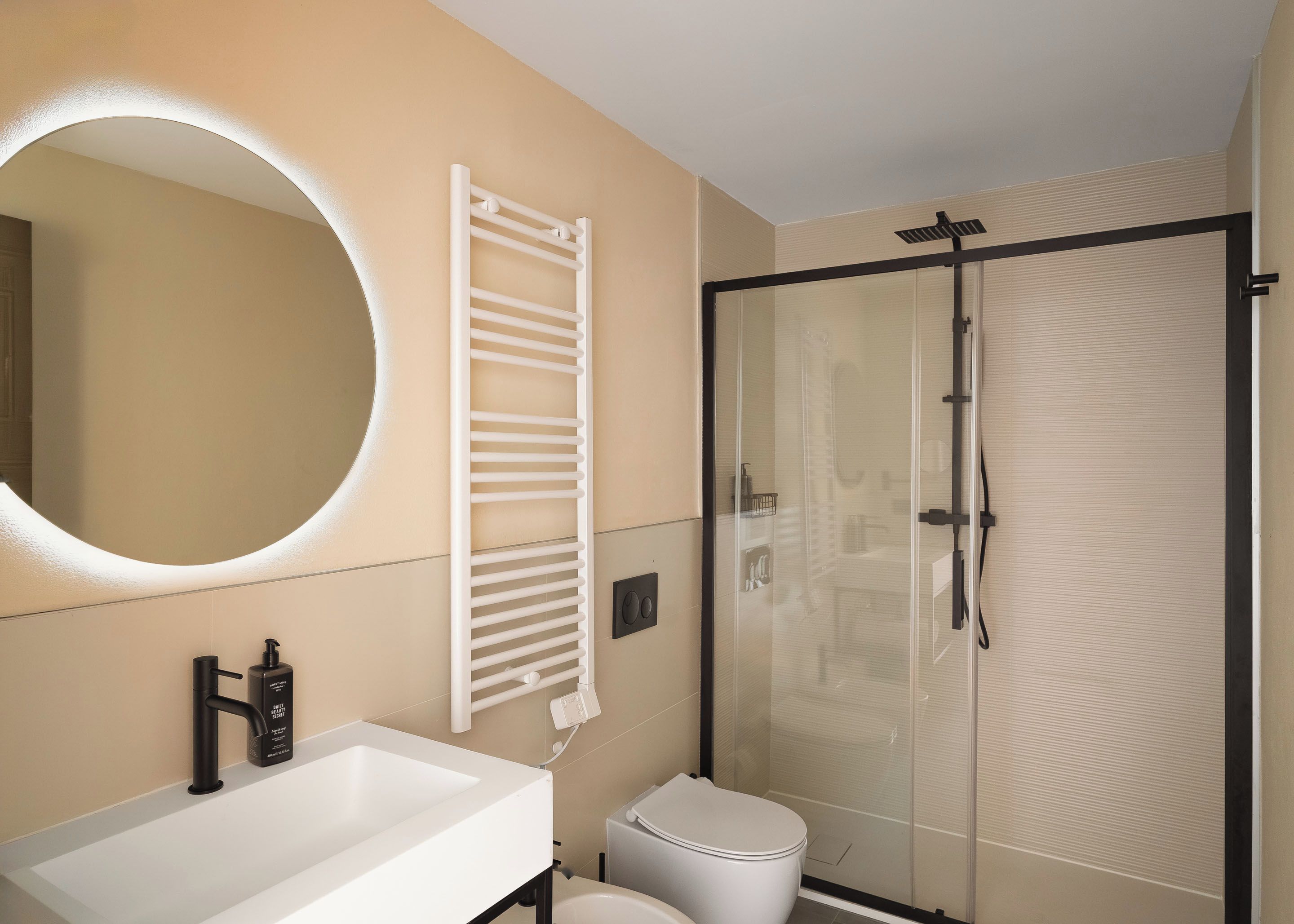 Ein modernes Badezimmer mit einem weißen Waschbecken, einem runden Spiegel, einem Handtuchhalter, einer Toilette und einer Duschkabine aus Glas.