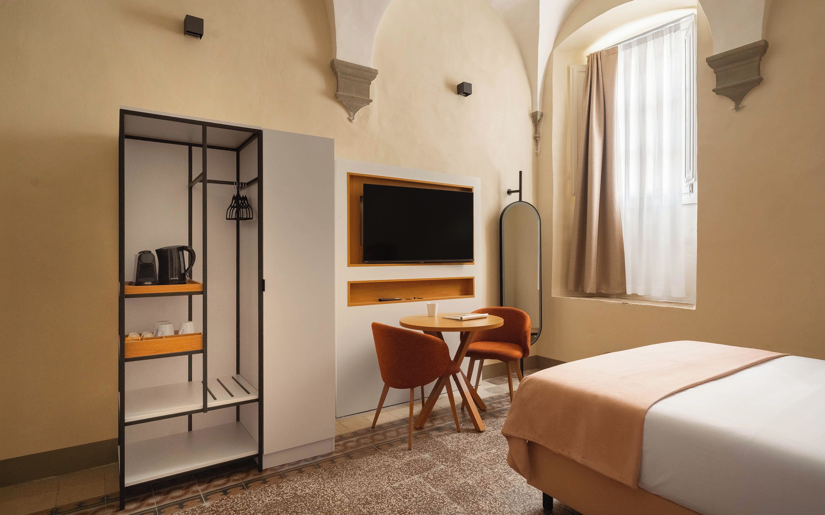 Una camera d'albergo con un letto, una scrivania con una sedia, un armadio, una televisione e una finestra con tende.