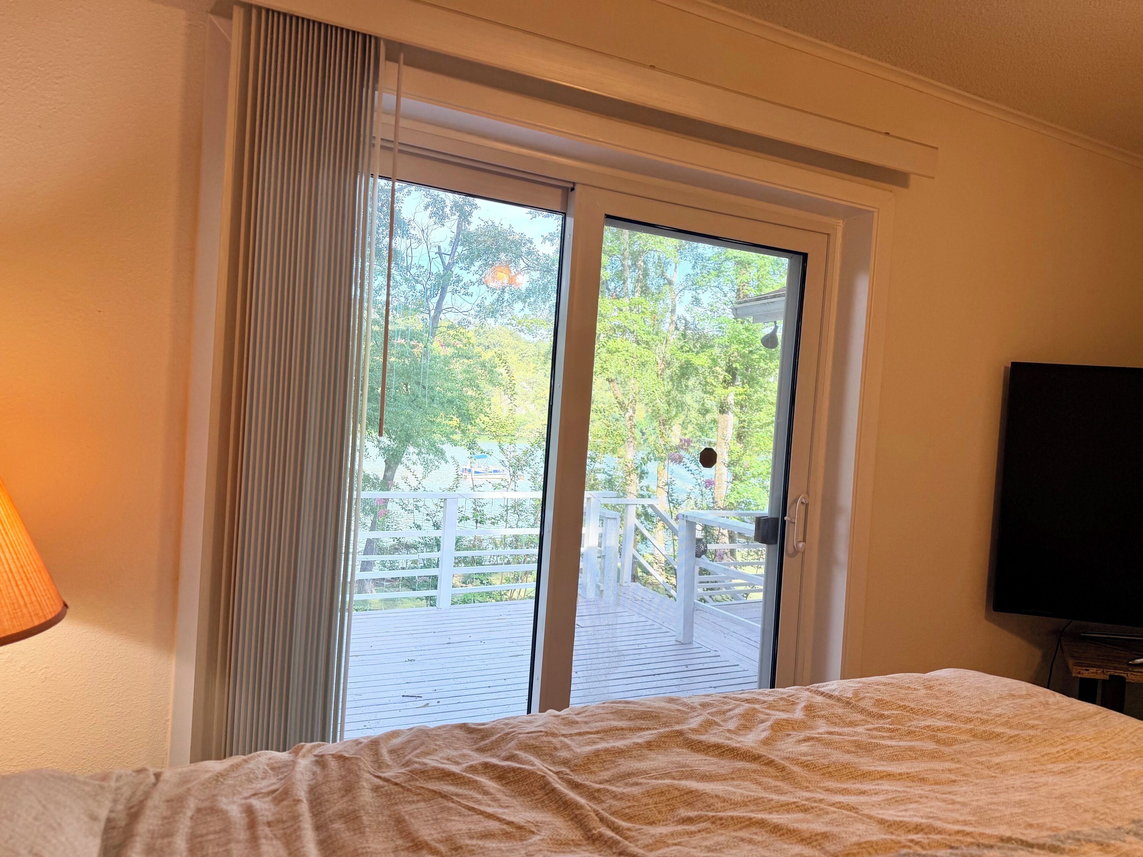 A hotel room with a bed, a television, a window with a view of a deck and trees, and a curtain.