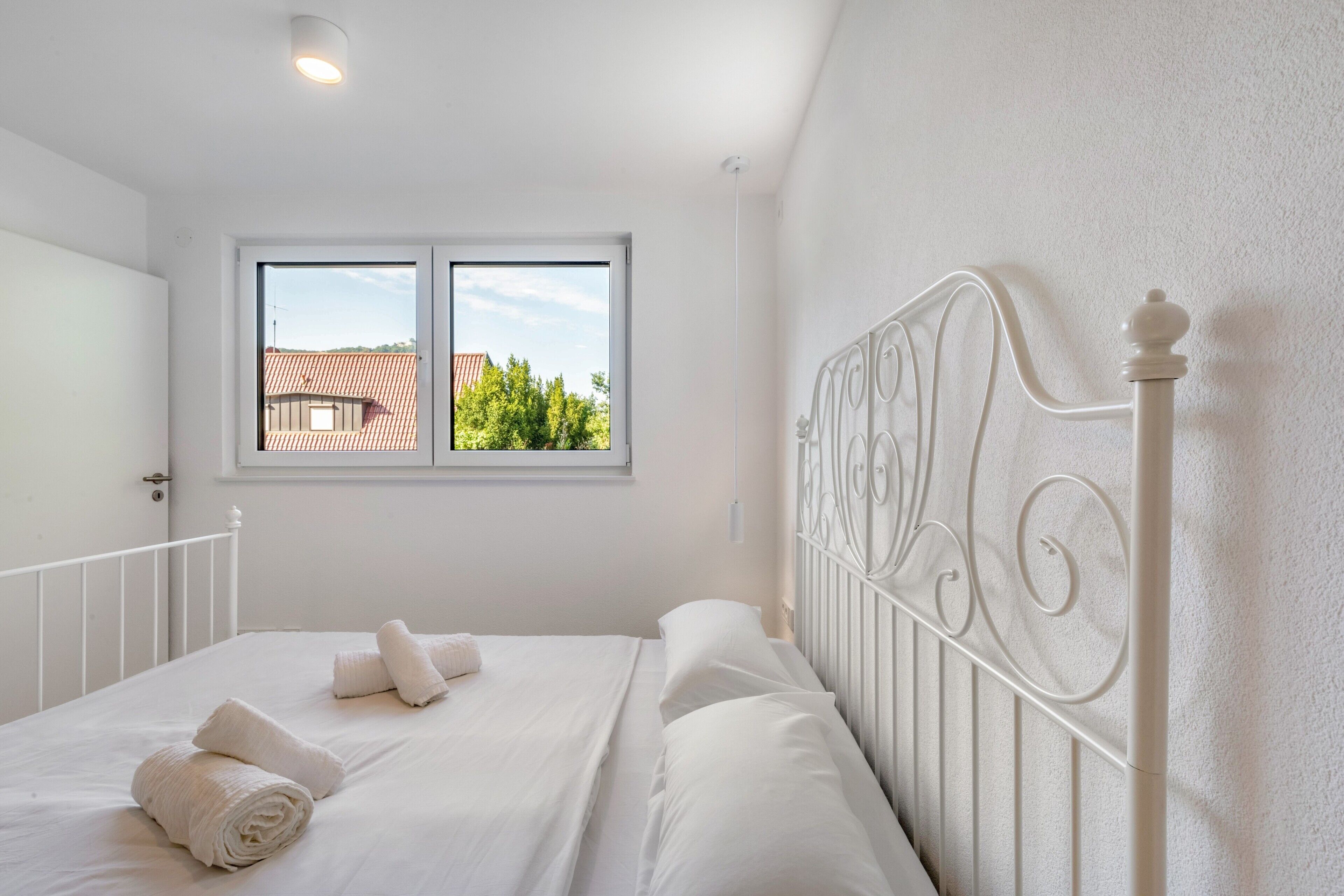 A bedroom with a white bed, a window with a view of a red-roofed building, and a white metal headboard.