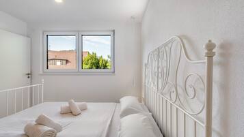 A bedroom with a white bed, a window with a view of a red-roofed building, and a white metal headboard.