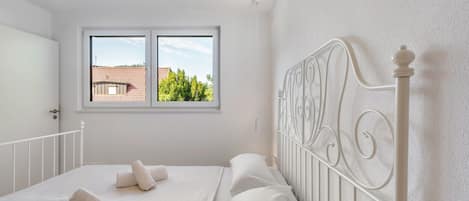 A bedroom with a white bed, a window with a view of a red-roofed building, and a white metal headboard.