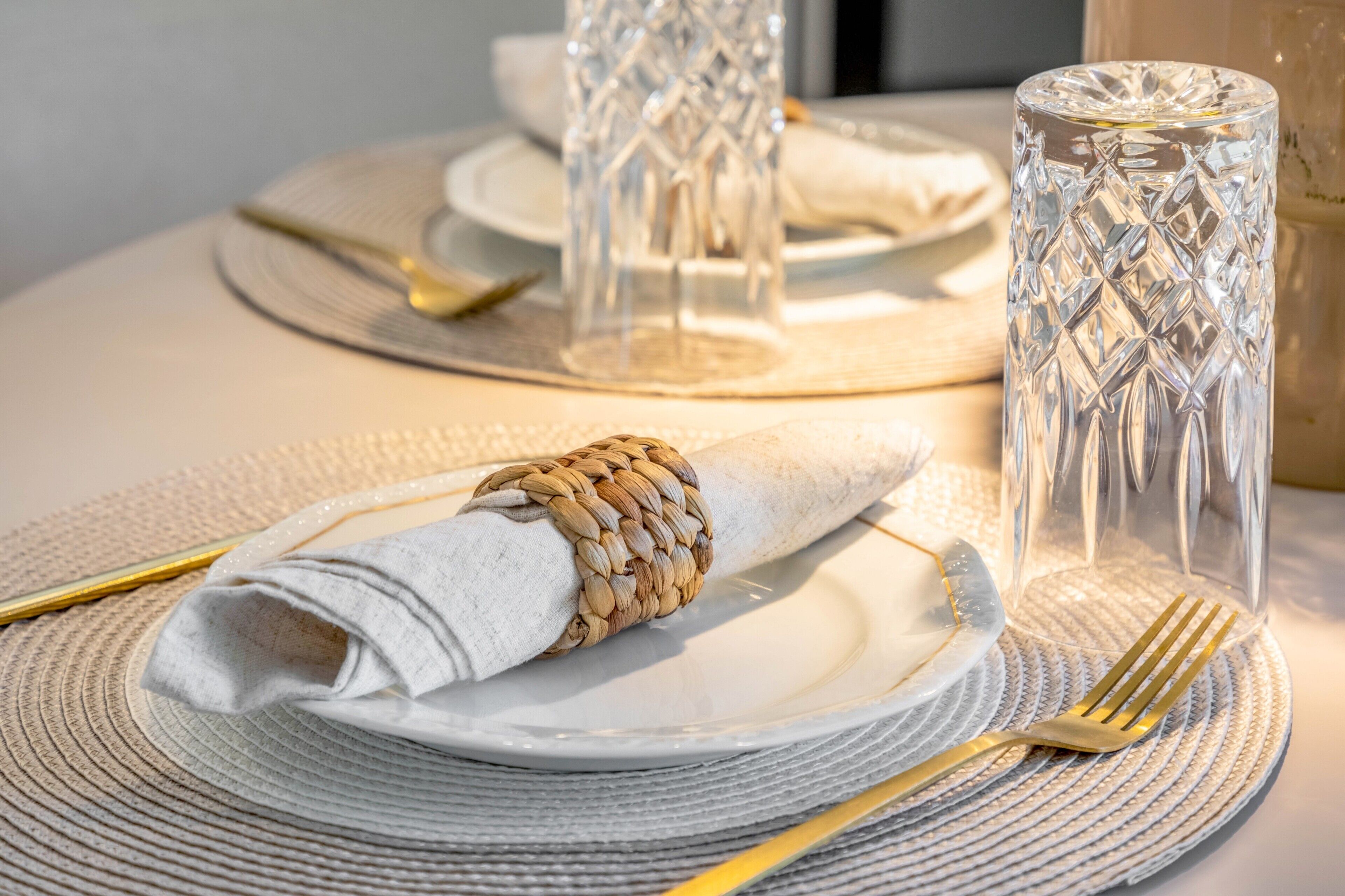 Une table dressée avec une serviette blanche, un porte-serviette en osier, un verre transparent, une fourchette dorée et une cuillère dorée.
