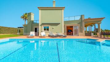 A modern house with a swimming pool, a patio with lounge chairs, and a clear blue sky.