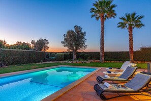 Une piscine rectangulaire dont le bord est revêtu de carreaux, entourée d’un espace patio aménagé avec des chaises longues et une table.