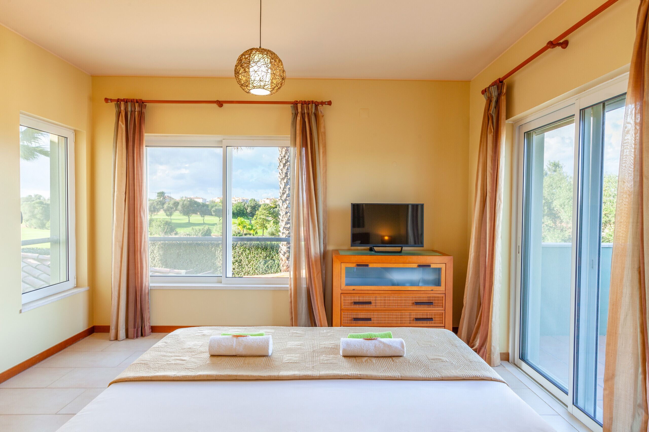A bedroom with a bed, a TV, a wooden dresser, and large windows with curtains.