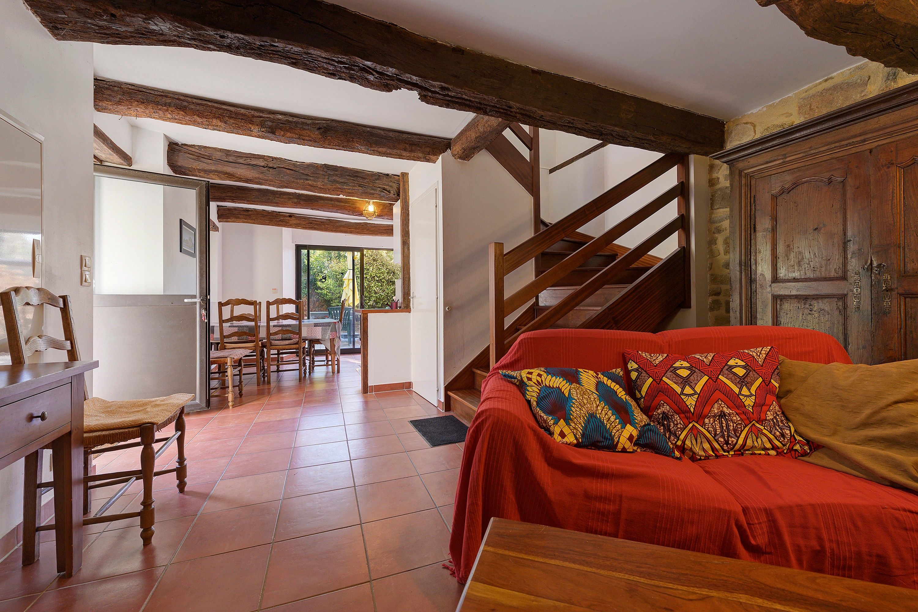 A room with a wooden staircase, a red sofa with patterned cushions, and a dining area with chairs and a table.