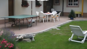 Een terras met een tafeltennistafel, witte stoelen en een witte parasol.