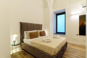 A neatly made bed with a headboard, a bedside table with a lamp, and a window with a view. - Charming suites nestled among art, the historic center, and the sea (Catania)