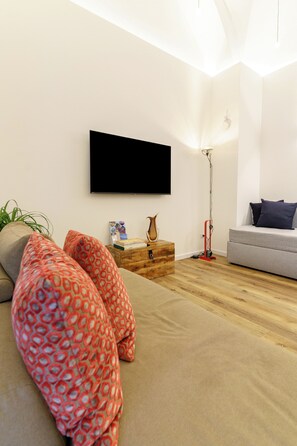 A modern living room with a flat-screen TV mounted on the wall, a grey sofa, a wooden side table with books and a vase, and a red floor lamp. - Charming suites nestled among art, the historic center, and the sea (Catania)