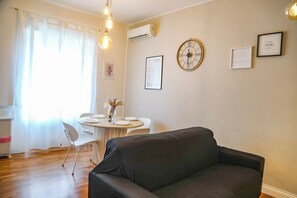 Un soggiorno con un divano scuro, un tavolo da pranzo rotondo con sedie e una sveglia a muro.