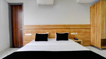 A hotel room with a bed, a wooden headboard, a nightstand with a phone, and an air conditioning unit.