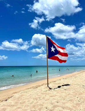 A beach with a flag on a pole, clear blue sky, and ocean. - Stay Car-Free in the Heart of Condado (San Juan)