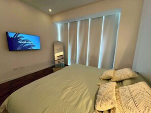 A bedroom with a large bed, a flat-screen TV mounted on the wall, and a window with vertical blinds. - Caracas VIP: 2H/2B + Fibra Thunder Net (Caracas)