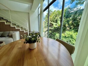 A modern interior with a wooden dining table, a potted plant, and a view of lush greenery through large windows. - Caracas VIP: 2H/2B + Fibra Thunder Net (Caracas)