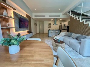 A modern living room with a wooden dining table, a grey sofa, and a staircase.