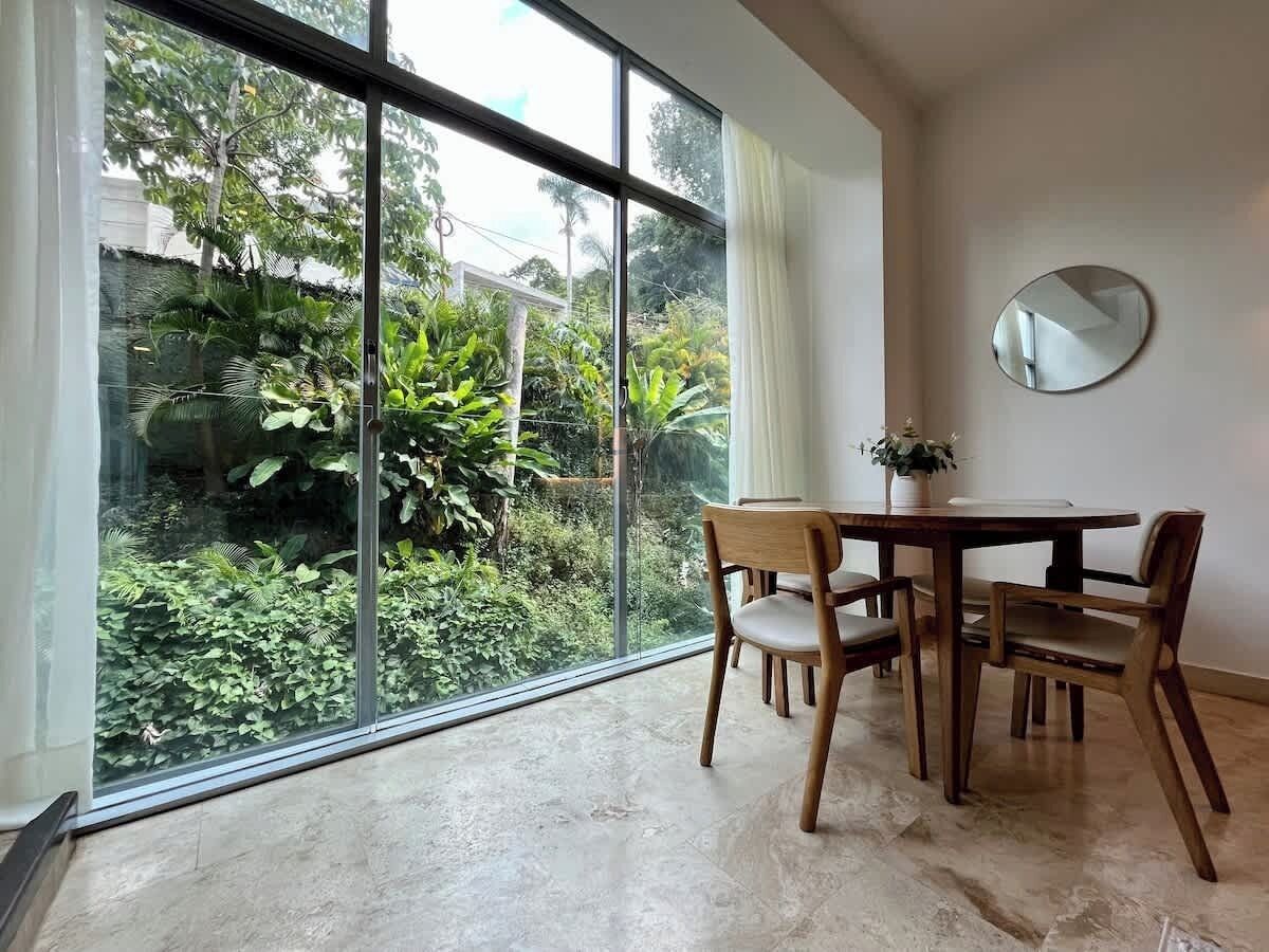 Un comedor con una mesa y sillas, un espejo redondo en la pared, y vistas a zonas verdes a través de amplios ventanales.