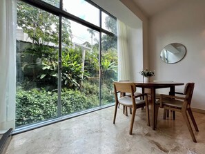 A dining area with a table and chairs, a round mirror on the wall, and a view of greenery through large windows.