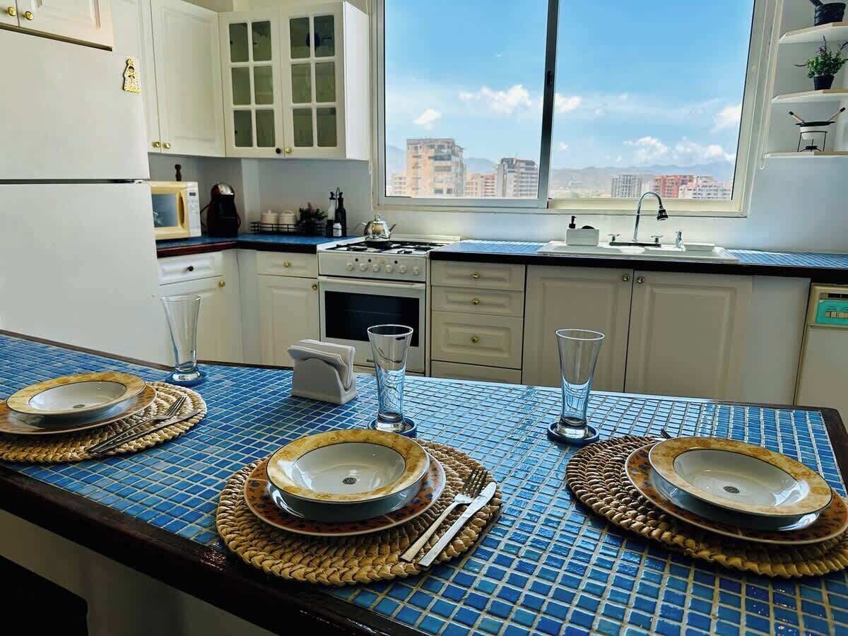 Een keuken met een blauwe betegelde achterwand, witte kastjes, een fornuis en een raam met uitzicht op de stad.
