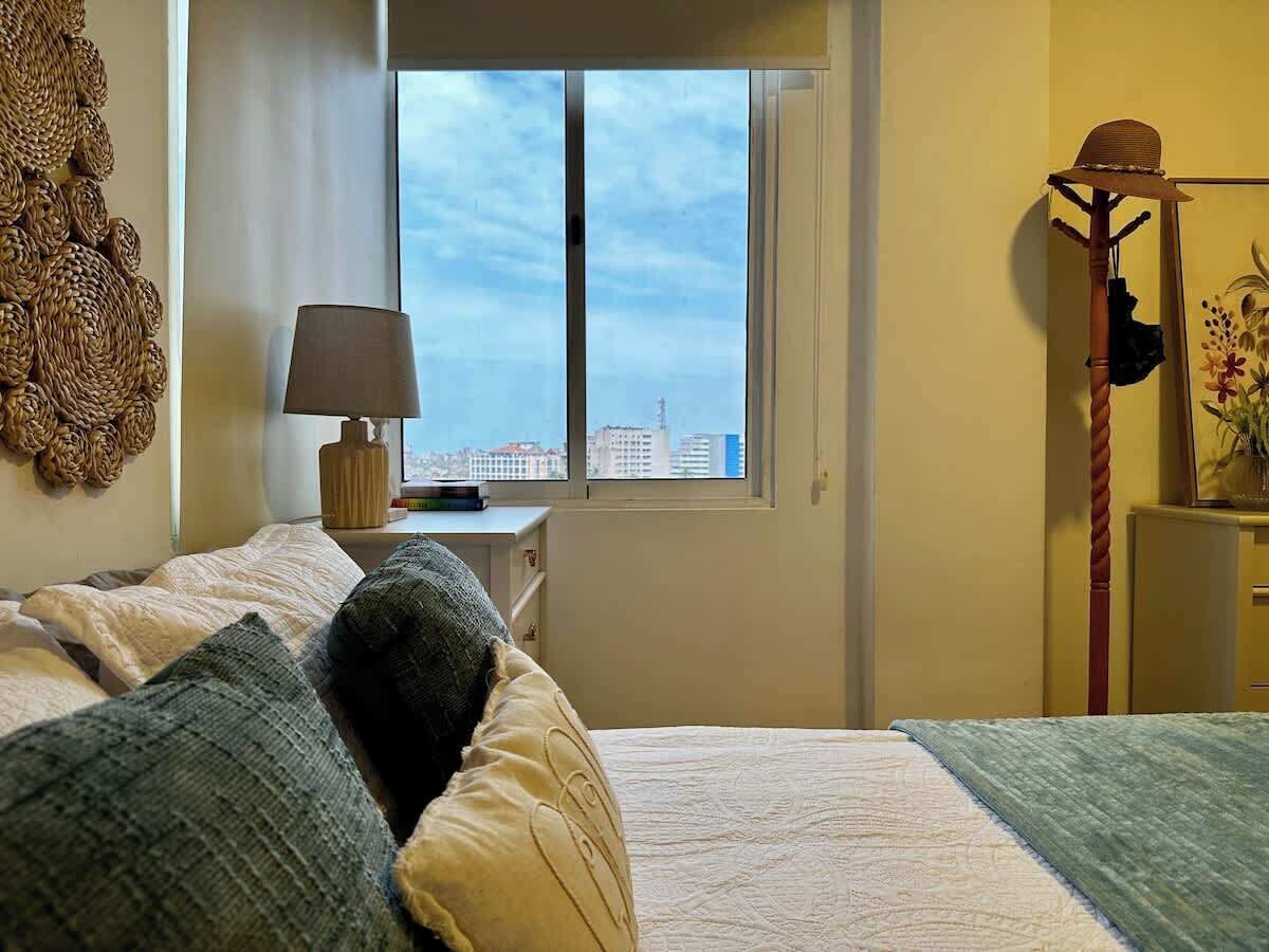Une chambre à coucher avec un lit, une table de chevet avec une lampe, une fenêtre donnant sur des immeubles et un élément décoratif mural.