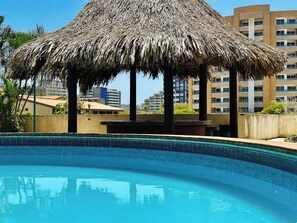 Ein Pool mit überdachter, reetgedeckter Struktur, umgeben von Gebäuden.