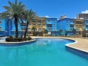 Uma área de piscina com borda curva, palmeiras e uma fileira de edifícios coloridos ao fundo.