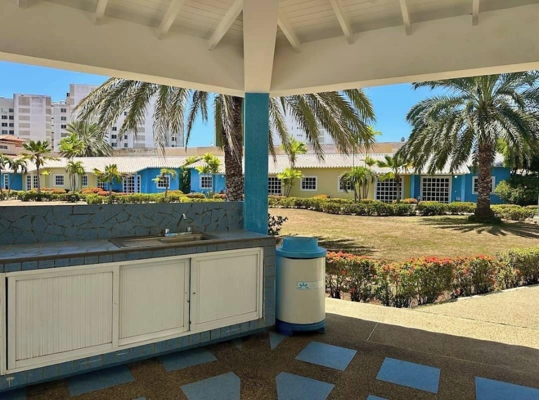 A covered outdoor area with a sink and trash bin, surrounded by palm trees and a garden.