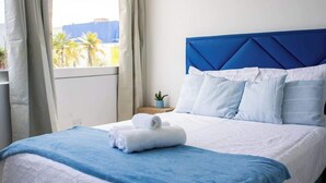 Een hotelkamer met een bed, nachtkastje, raam met gordijnen en uitzicht op palmbomen.