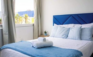 Ein Hotelzimmer mit einem Bett, einem Nachttisch, einem Fenster mit Vorhängen und Blick auf Palmen.