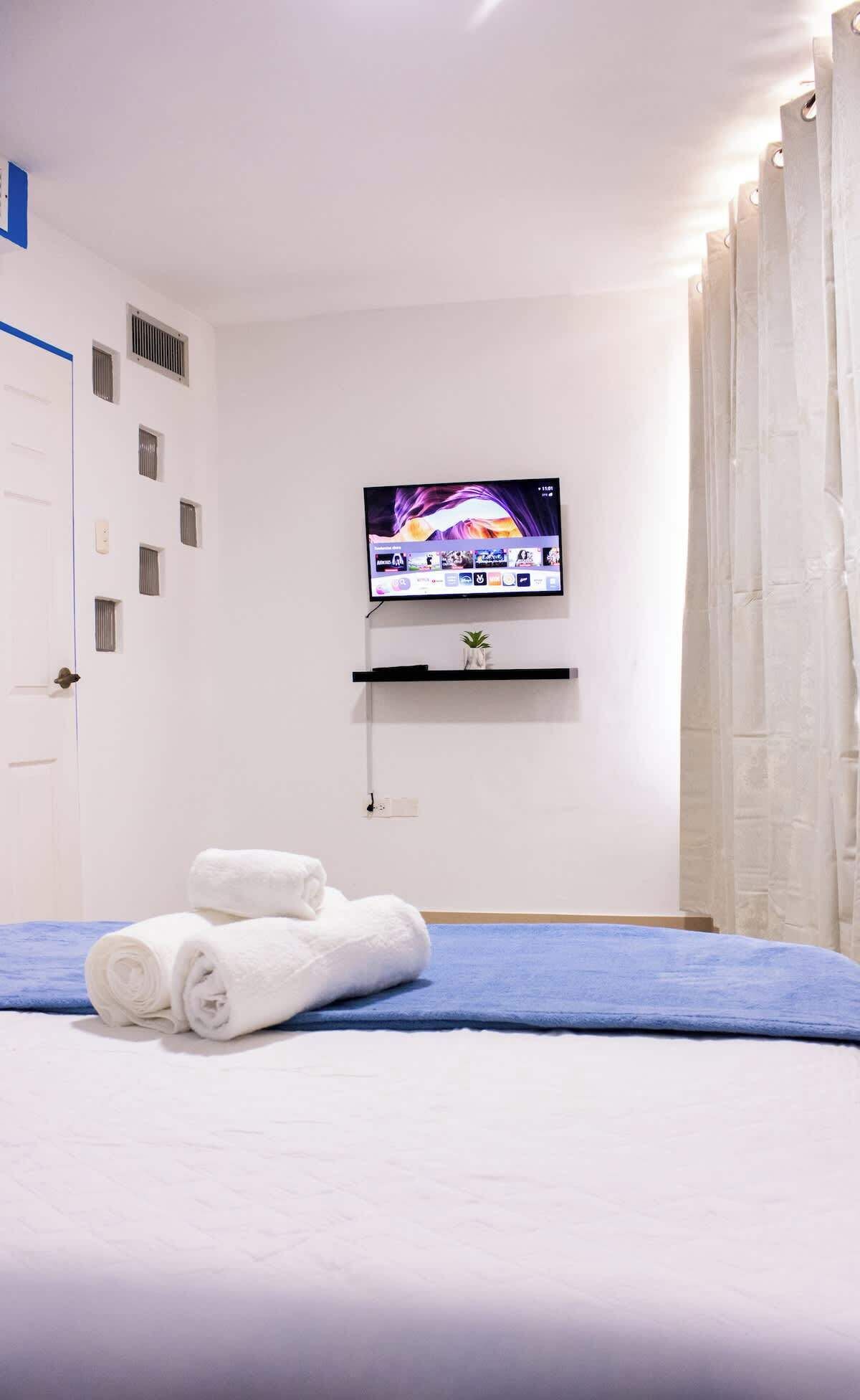 Une chambre d’hôtel avec un lit, un téléviseur fixé au mur, une porte avec une bande bleue et une fenêtre avec des rideaux.