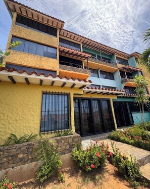 Un edificio de varios pisos con balcones, ventanas grandes y un jardín con plantas y flores.