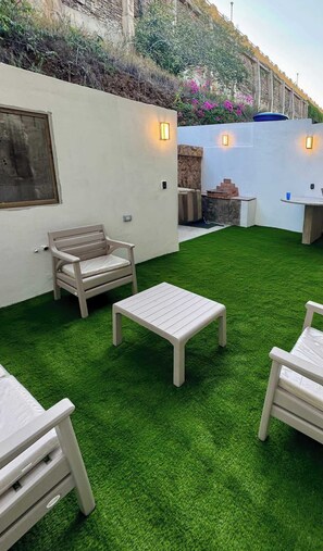 Eine Terrasse im Freien mit Kunstgrashalm, einem weißen Tisch und zwei weißen Stühlen.