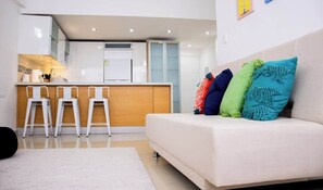 A modern living room with a sofa and colorful cushions, a kitchen area with a bar, and a white floor. - Ocean Blue/ Vista y Atardeceres (Porlamar)