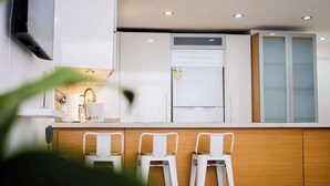 Eine moderne Küche mit weißen Schränken, einem eingebauten Ofen und einem Kühlschrank. Vor der Arbeitsplatte stehen drei weiße Barhocker.