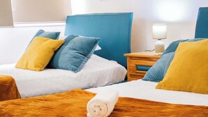 Ein Hotelzimmer mit zwei Betten, jeweils mit einem blauen Kopfteil, einem Nachttisch aus Holz mit einer Lampe und einem zusammengerollten Handtuch auf dem Bett.