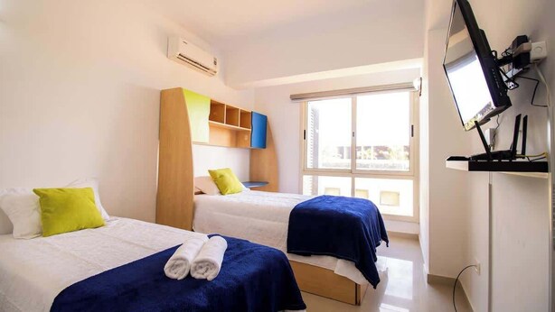 Een hotelkamer met twee bedden, een aan de muur bevestigde televisie en een raam dat natuurlijk licht biedt.
