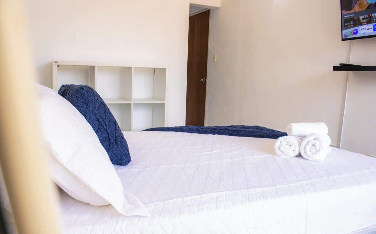 Habitación de hotel con cama, cabecera blanca, televisor montado en la pared y puerta que da a otra habitación.
