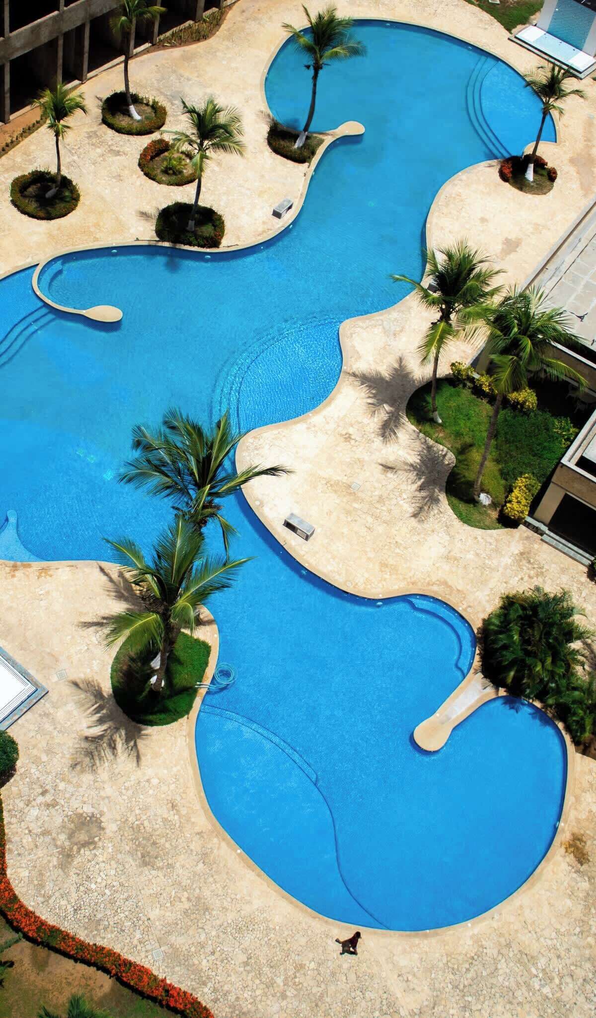 Una grande piscina sinuosa con diverse sezioni, circondata da palme e da una zona sabbiosa.