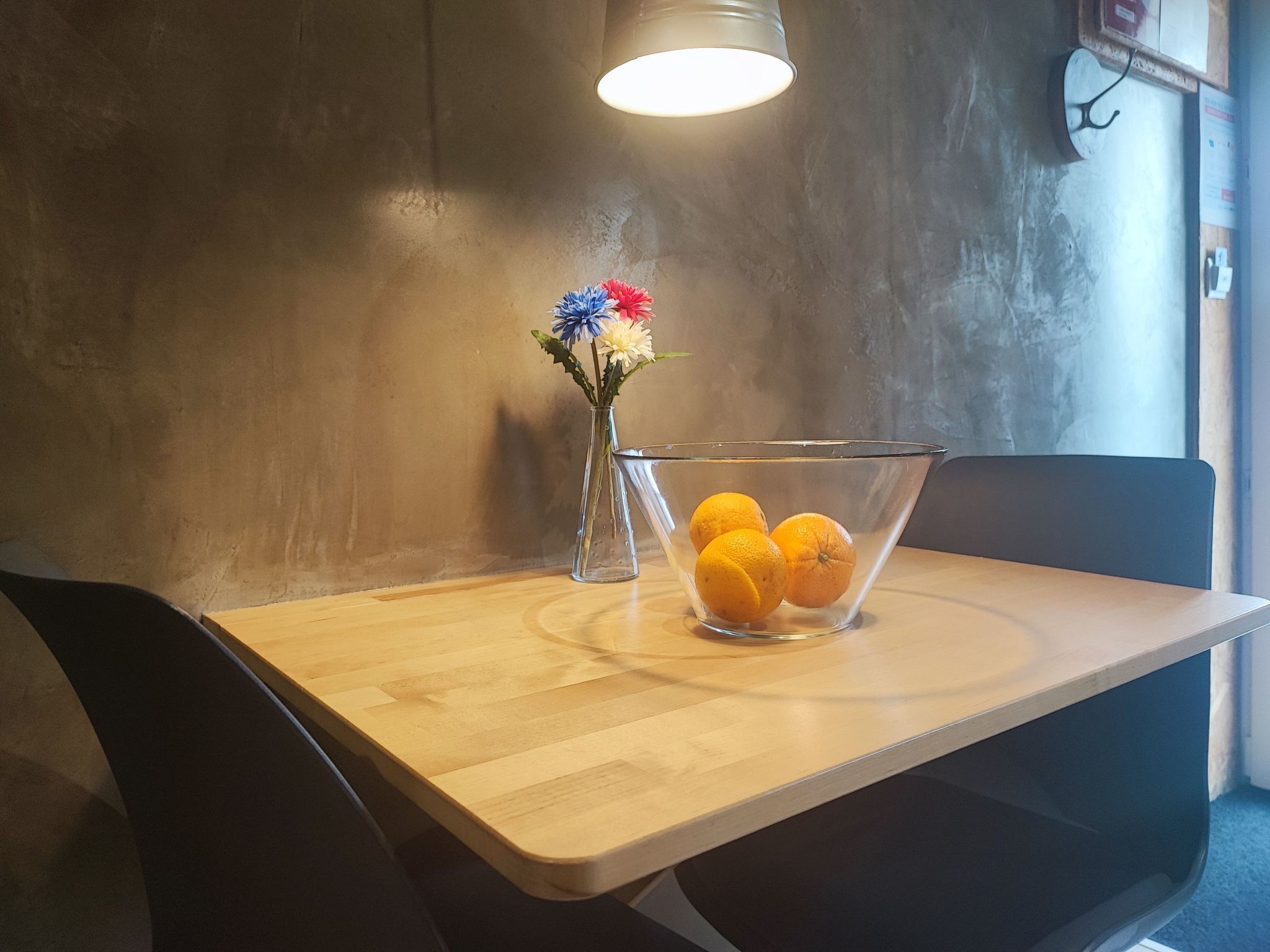 Uma mesa de madeira com uma tigela de vidro transparente cheia de laranjas, um vaso com flores e um candeeiro pendente.