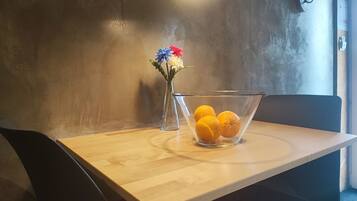 Uma mesa de madeira com uma tigela de vidro transparente cheia de laranjas, um vaso com flores e um candeeiro pendente.